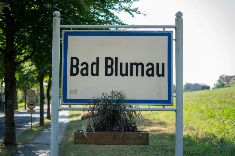 A white sign with blue borders and black text reads 'Bad Blumau'. Below the sign, a wooden planter with greenery is attached to the bottom.