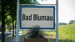 A white sign with blue borders and black text reads 'Bad Blumau'. Below the sign, a wooden planter with greenery is attached to the bottom.