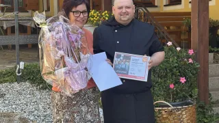 A man and a woman stand outside a house, the man holding a certificate and flowers, smiling for the camera.