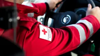 A person in a red uniform with a red cross patch on the left arm is driving a car.