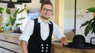 A man with glasses and a vest stands in front of a wooden counter, smiling at the camera.