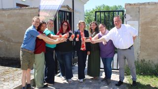 A group of people, both men and women, are standing outside and holding glasses of orange juice. They are smiling and appear to be celebrating. Behind them is a banner and a black gate.