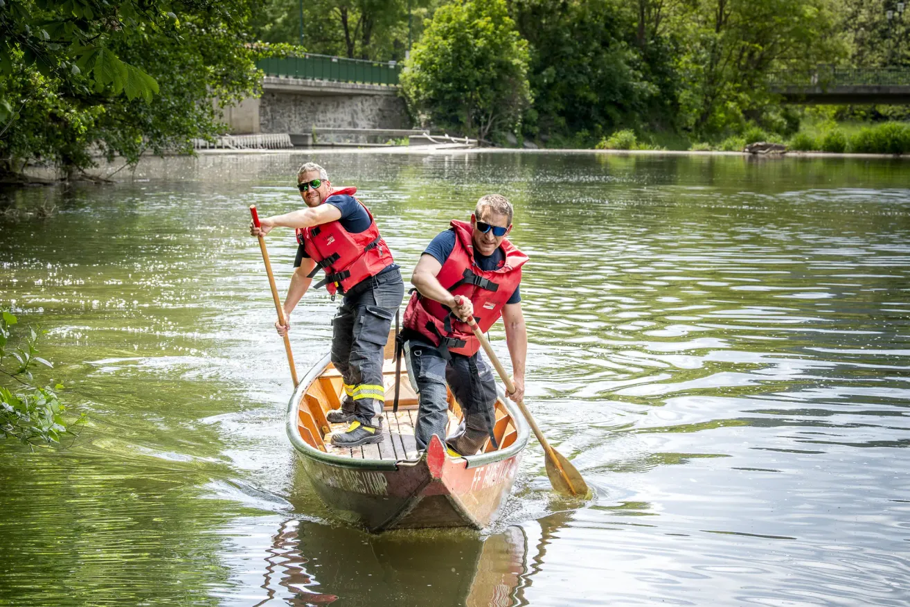 Two men in life jackets paddle a canoe on a calm river, with a bridge and trees in the background.