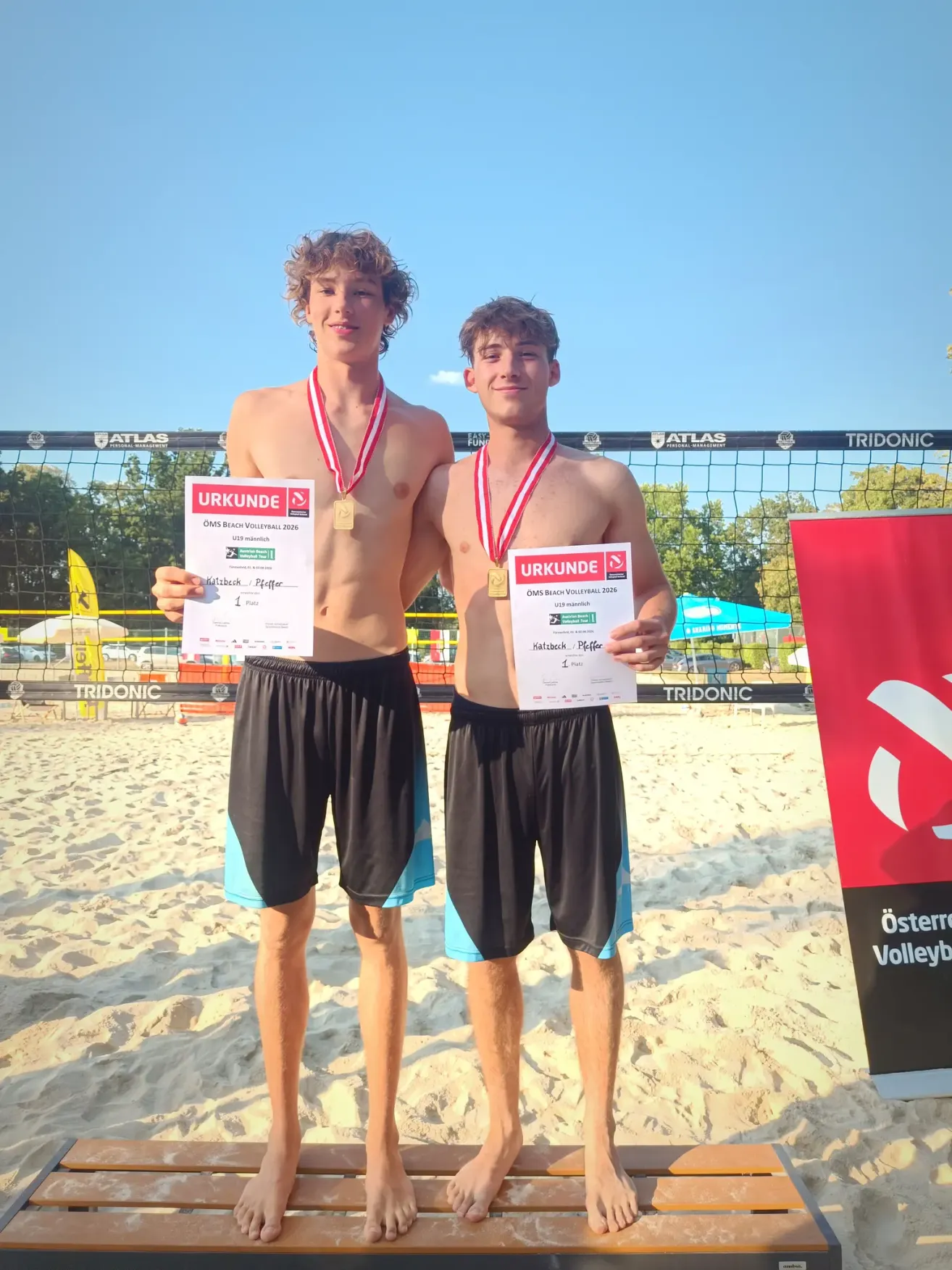 Zwei junge Männer stehen auf einem Strand, beide halten Medaillen und Urkunden. Sie tragen schwarze Shorts mit blauen Besätzen. Hinter ihnen steht ein Volleyballnetz mit einem Banner, das 'TRIDONIC' besagt.