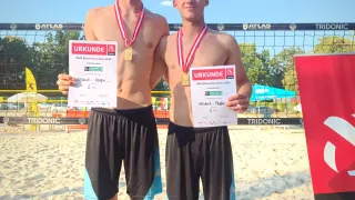 Zwei junge Männer stehen auf einem Strand, beide halten Medaillen und Urkunden. Sie tragen schwarze Shorts mit blauen Besätzen. Hinter ihnen steht ein Volleyballnetz mit einem Banner, das 'TRIDONIC' besagt.
