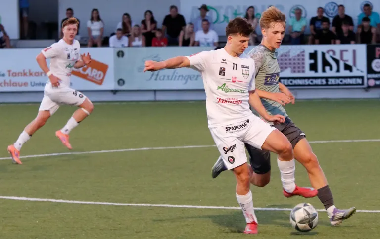 Two soccer players are competing on a field, one in a white jersey with the number 11, and the other in a gray jersey. They are both running towards the ball.