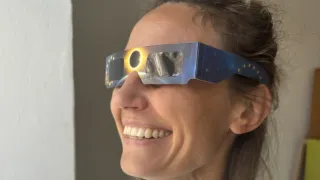 A woman wearing solar eclipse glasses, smiling with a view of the sun partially eclipsed through the glasses.