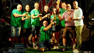 Eine Gruppe von Menschen in grünen Shirts und Shorts hält Gläser mit Bier vor einem roten und weißen Logo.