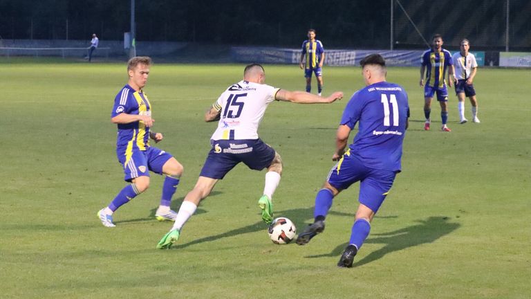Three soccer players compete on a field at night, one in a white jersey with the number 15, another in a blue jersey with the number 11, and a third in a blue and yellow jersey.
