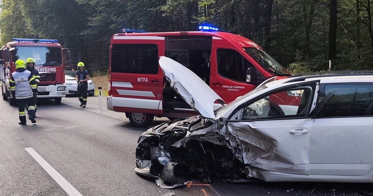A wrecked white car lies on the side of a road with its hood up. A red emergency vehicle with 'MTFA' on the side is parked nearby, and a person in a yellow helmet is standing beside a white car.
