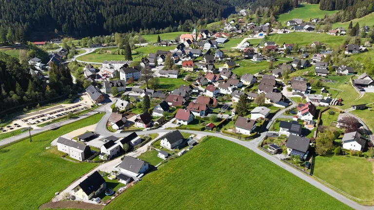Aerial view of a small village surrounded by green fields and forested hills. The village has a mix of traditional and modern houses, some with solar panels.