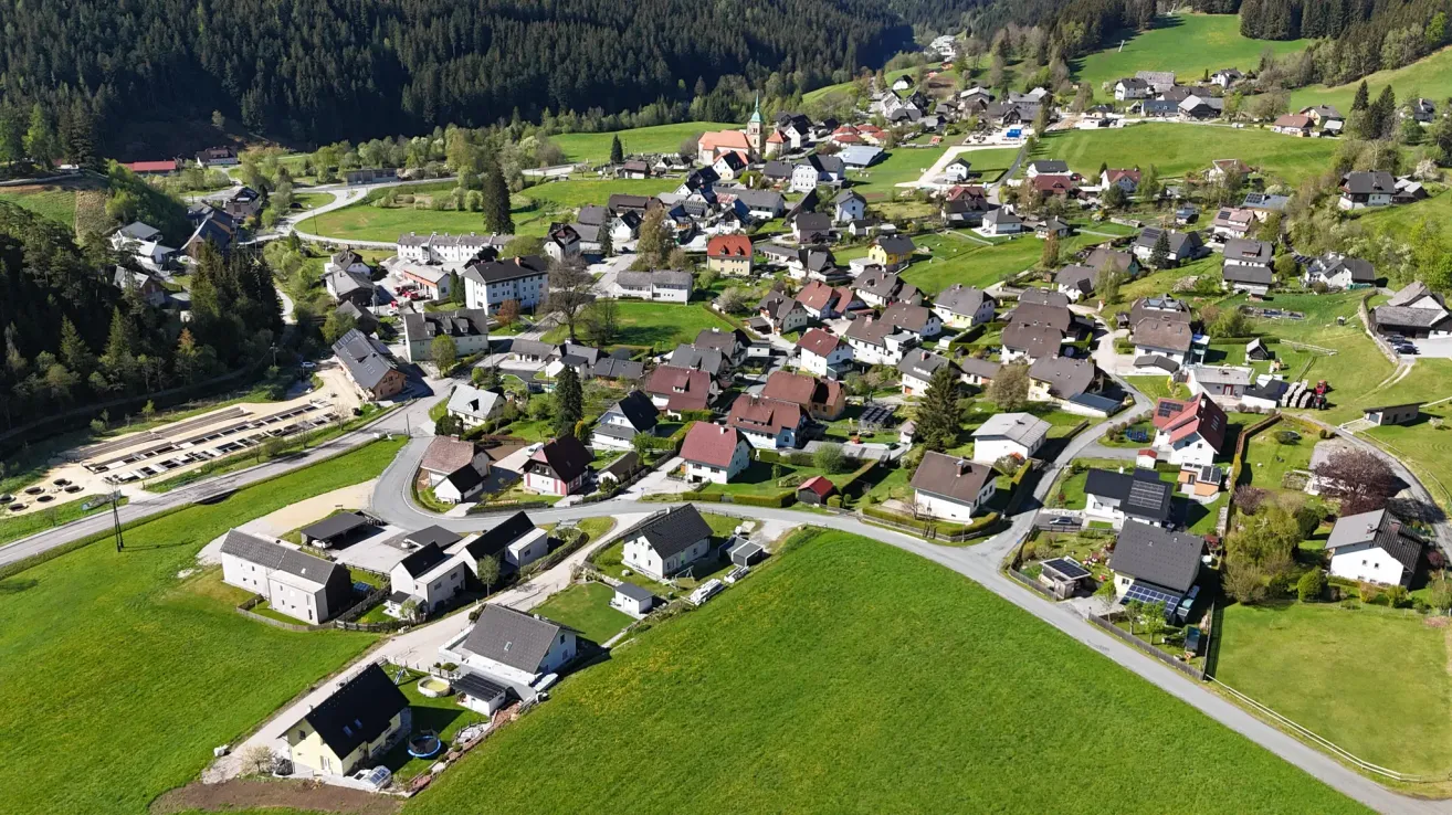 Aerial view of a small village surrounded by green fields and forested hills. The village has a mix of traditional and modern houses, some with solar panels.