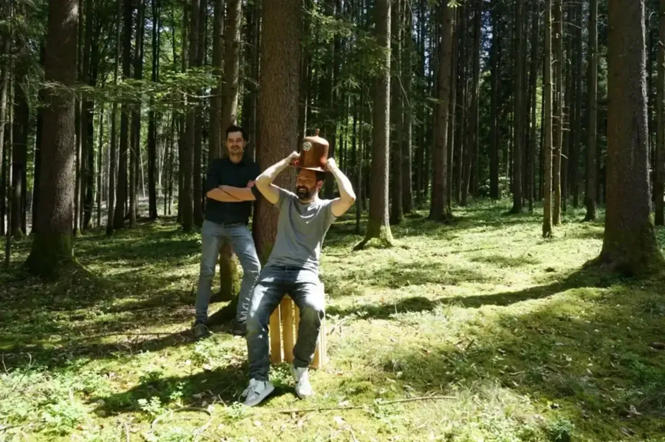 Two men in a forest, one sitting on a wooden stump, holding a wooden hat. The other stands behind, watching.