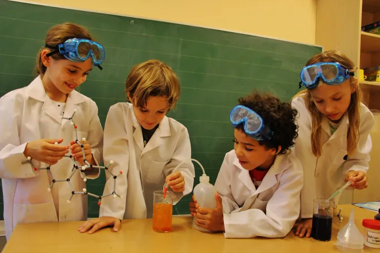 Four children in white lab coats and goggles are conducting a science experiment in a classroom. One child is holding a molecular model, another is pouring liquid into a beaker.