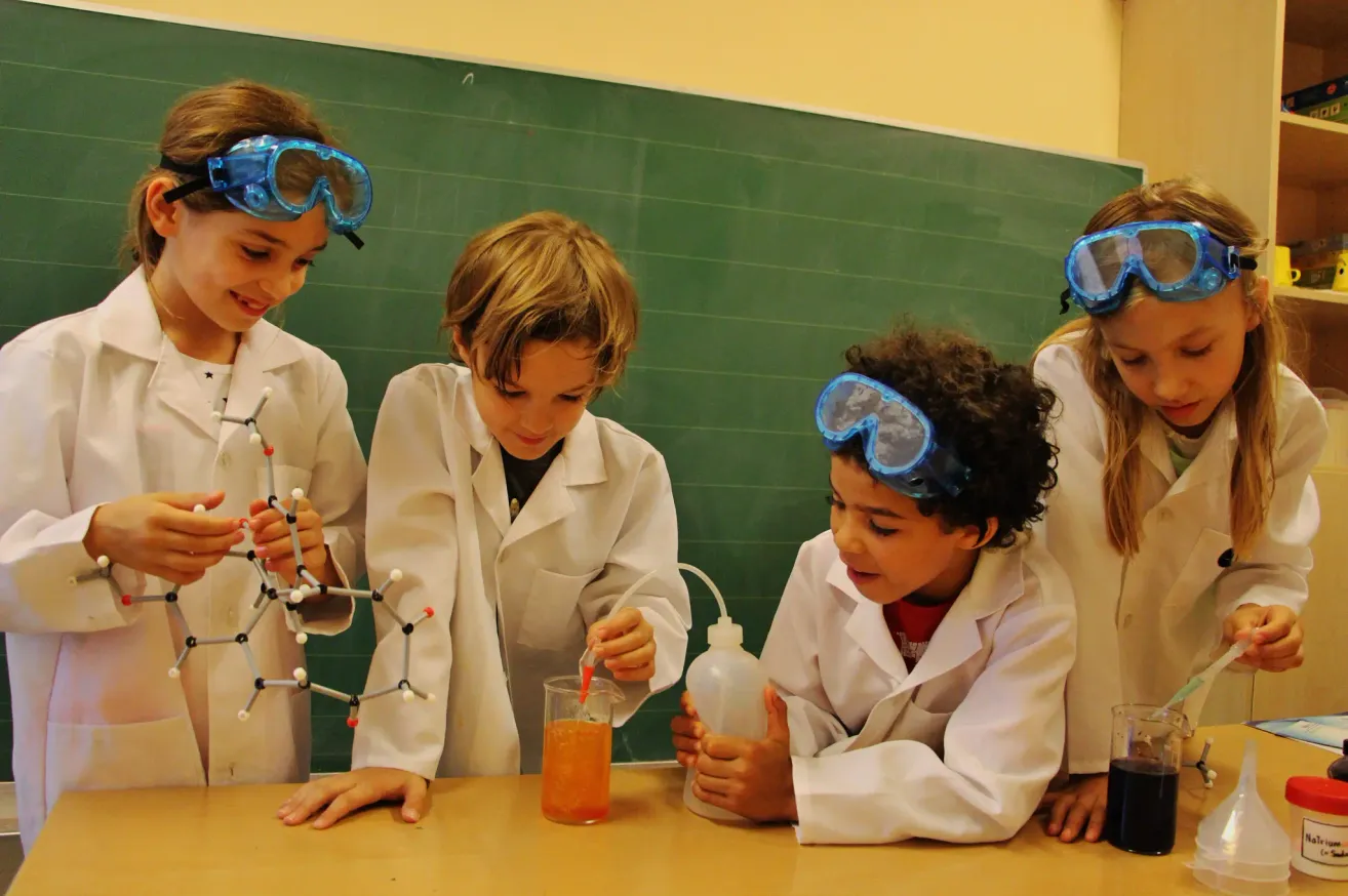 Four children in white lab coats and goggles are conducting a science experiment in a classroom. One child is holding a molecular model, another is pouring liquid into a beaker.