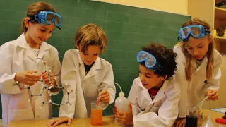 Four children in white lab coats and goggles are conducting a science experiment in a classroom. One child is holding a molecular model, another is pouring liquid into a beaker.