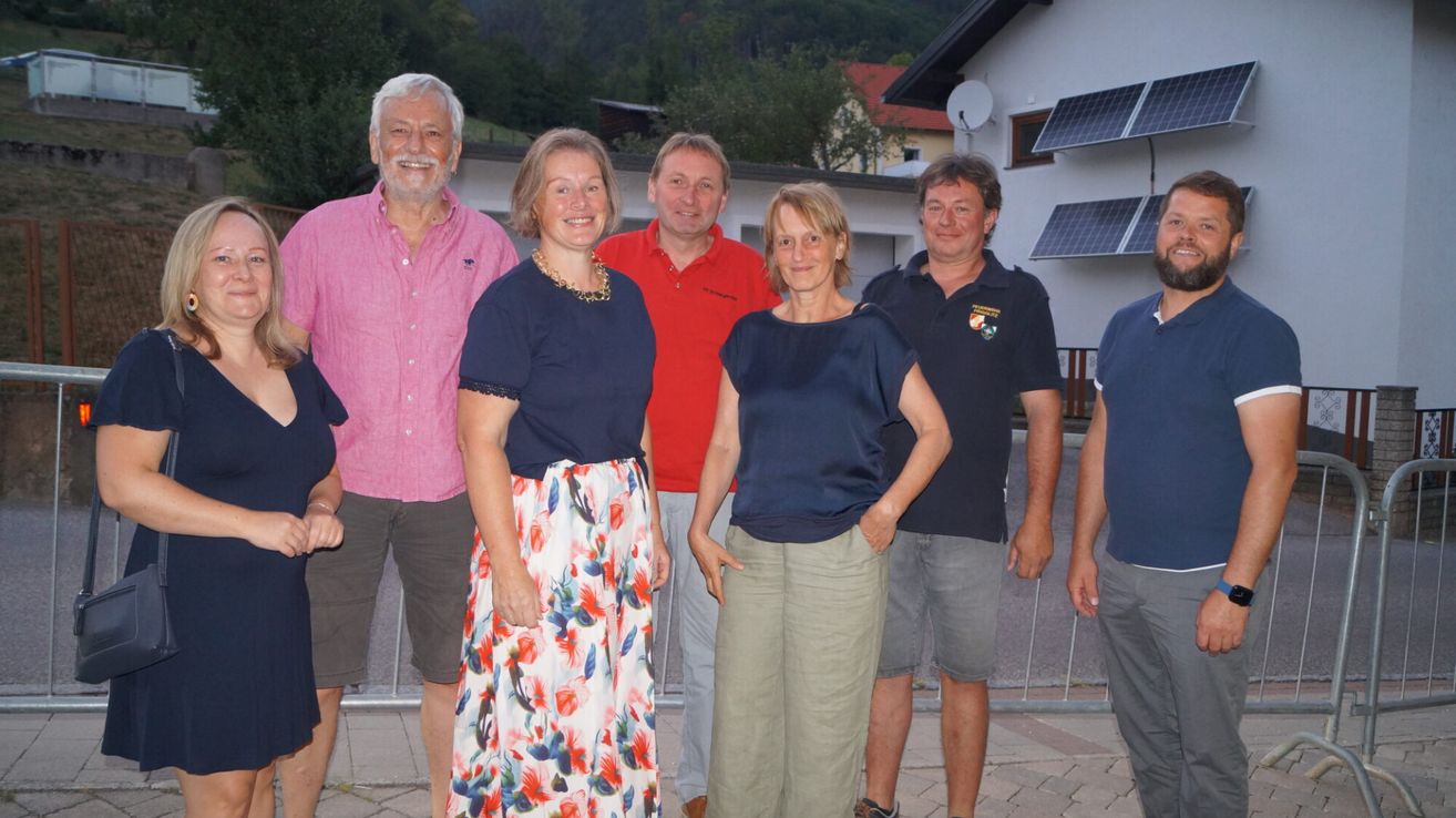 A group of six people, three men and three women, are standing together in front of a house with solar panels on the roof. They are smiling and appear to be posing for a photograph.
