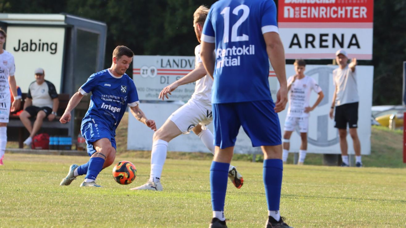 A soccer player in blue is chasing a ball while another player in white is defending. The player in blue has the number 12 on his jersey.