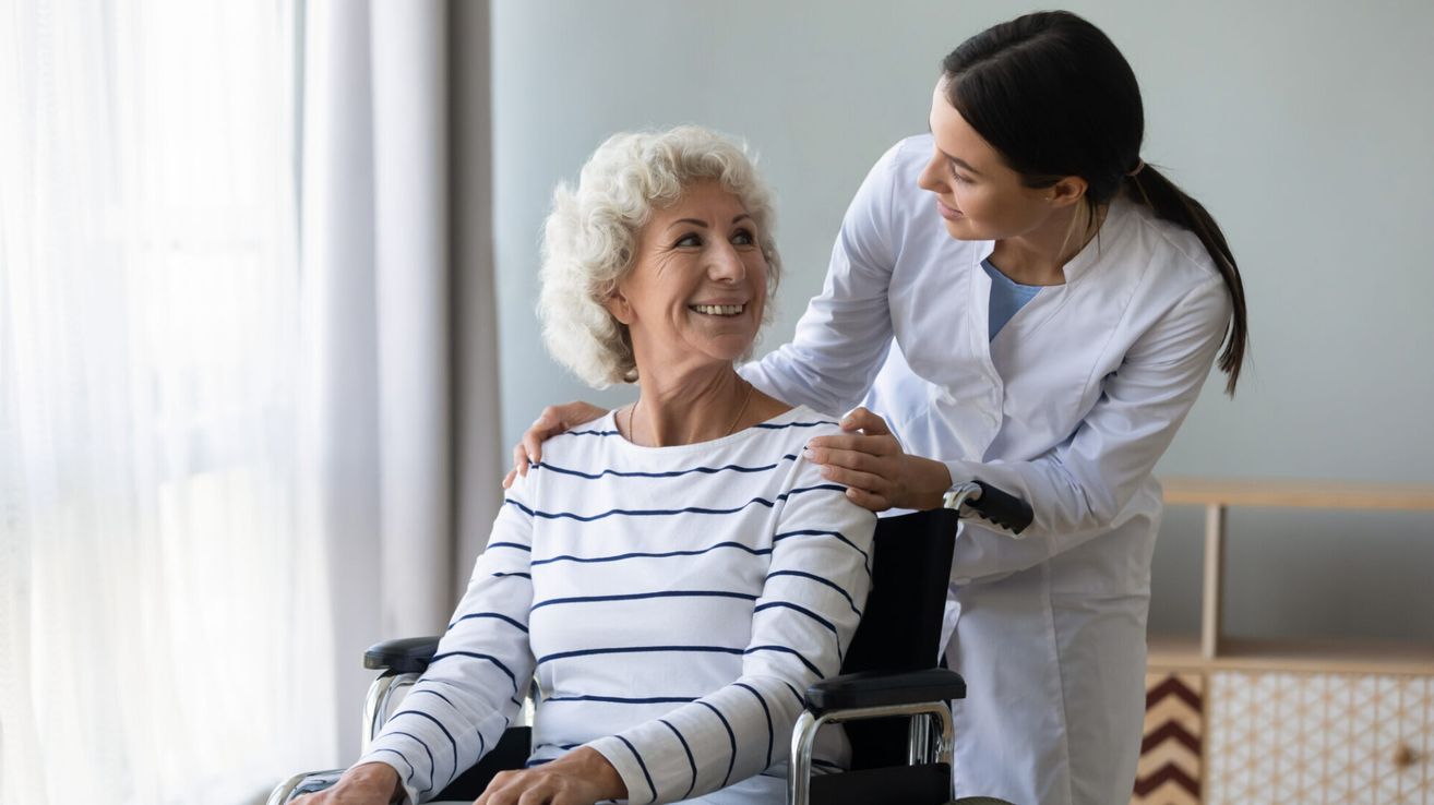 An elderly woman in a wheelchair smiles as a nurse stands beside her, offering support and a comforting presence.