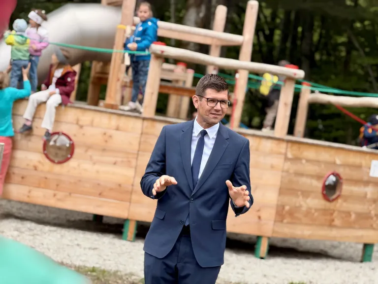 Ein Mann in einem blauen Anzug und mit Brille steht vor einer hölzernen Spielstruktur, lächelt und gestikuliert, während er spricht. Kinder spielen im Hintergrund auf der Struktur.