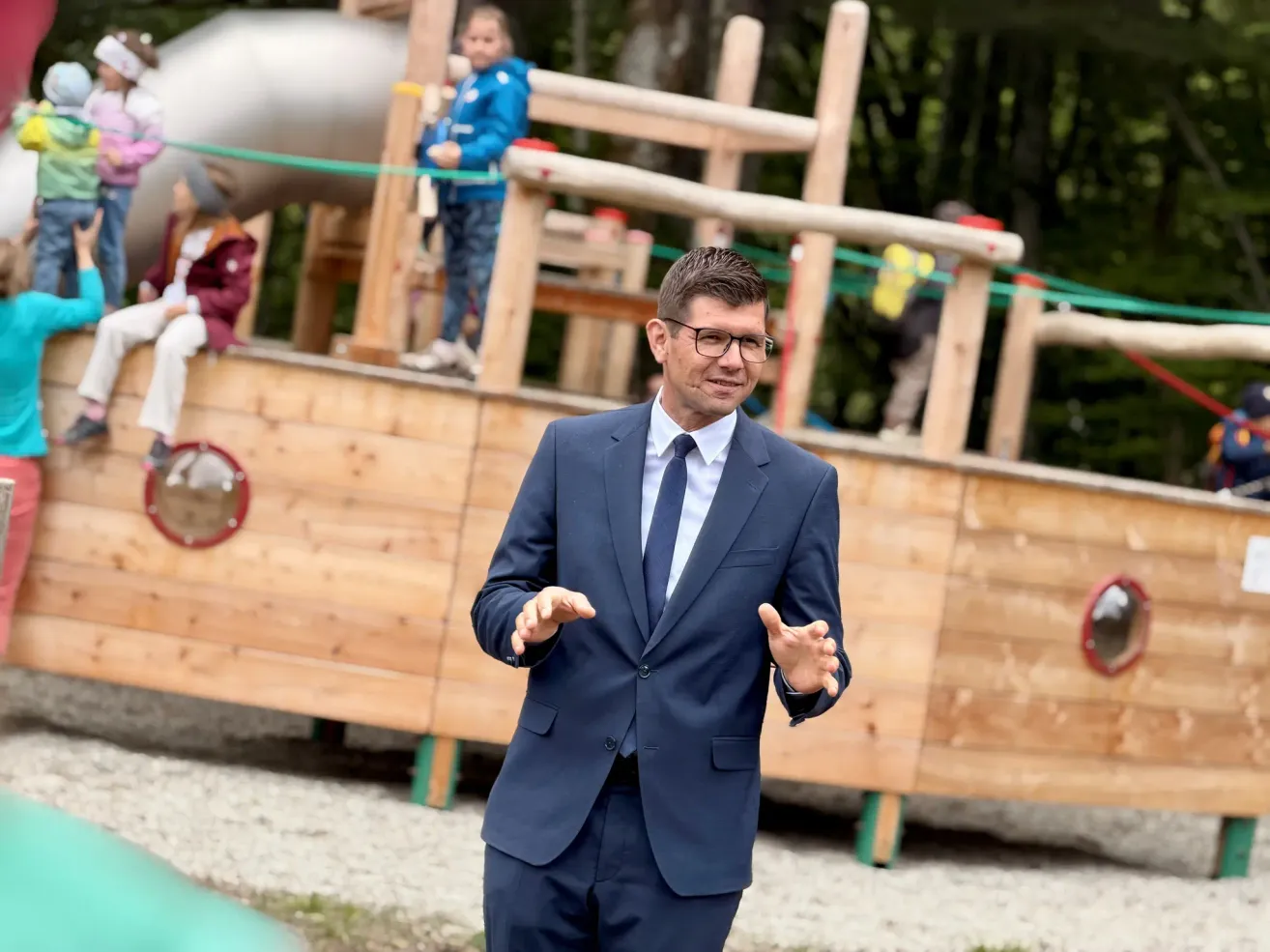 Ein Mann in einem blauen Anzug und mit Brille steht vor einer hölzernen Spielstruktur, lächelt und gestikuliert, während er spricht. Kinder spielen im Hintergrund auf der Struktur.