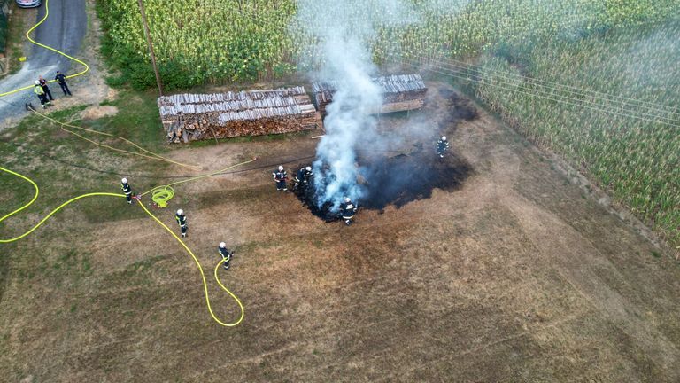Feuerwehrleute löschen einen Brand auf einem offenen Feld, mit Holzstapeln und einer Schlauch in der Nähe.