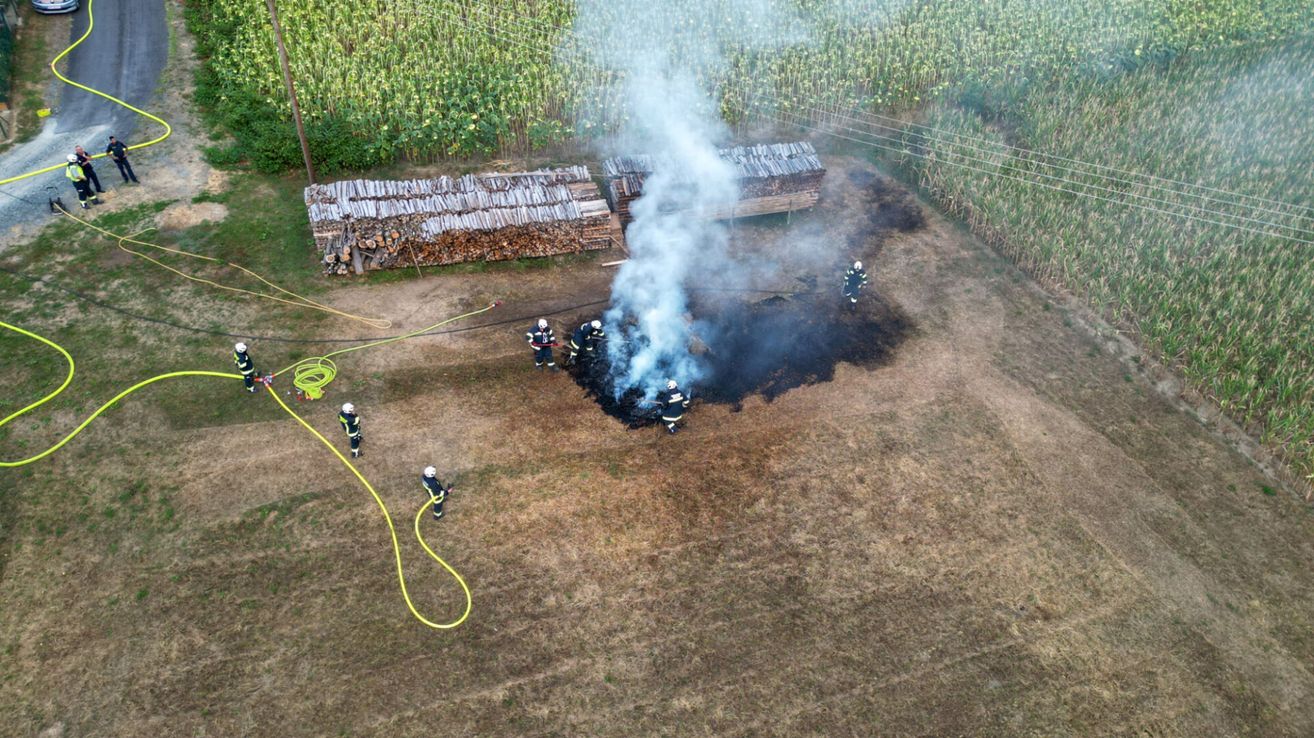 Feuerwehrleute löschen einen Brand auf einem offenen Feld, mit Holzstapeln und einer Schlauch in der Nähe.