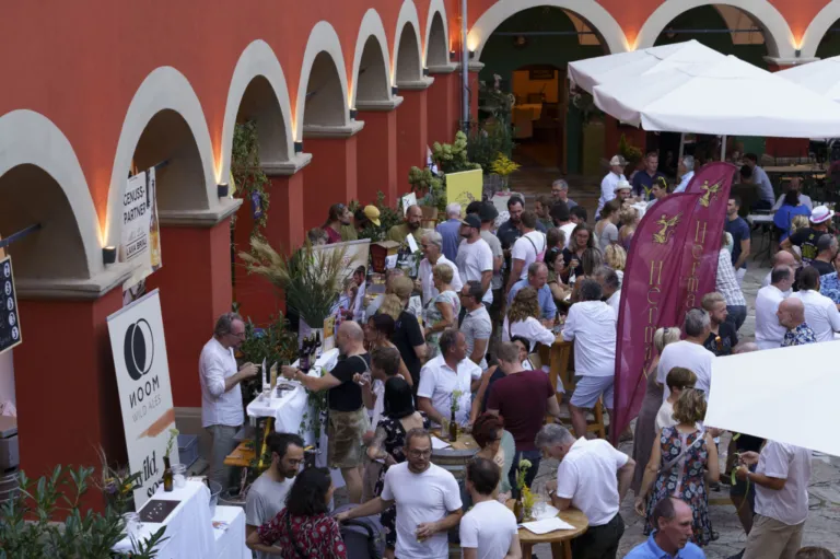A bustling outdoor event with people mingling around tables, some seated and others standing. The setting is adorned with plants, banners, and a red-walled building with arched openings.
