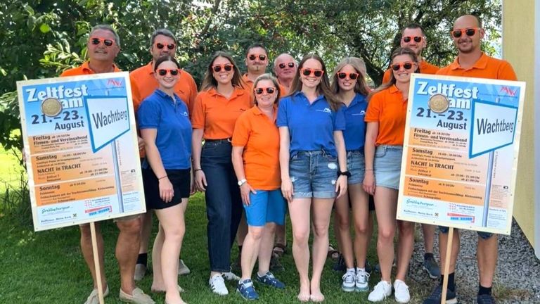 Eine Gruppe von Menschen in orangen und blauen Hemden posiert für ein Foto im Park. Sie tragen alle Sonnenbrillen.