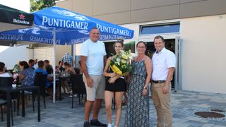 Vier Personen stehen vor einem Gebäude. Eine Frau hält einen Blumenstrauß. Der Mann links trägt ein weißes T-Shirt und Shorts. Der Mann rechts trägt ein weißes Hemd und Hose. Die Frau in der Mitte trägt ein schwarzes Kleid.