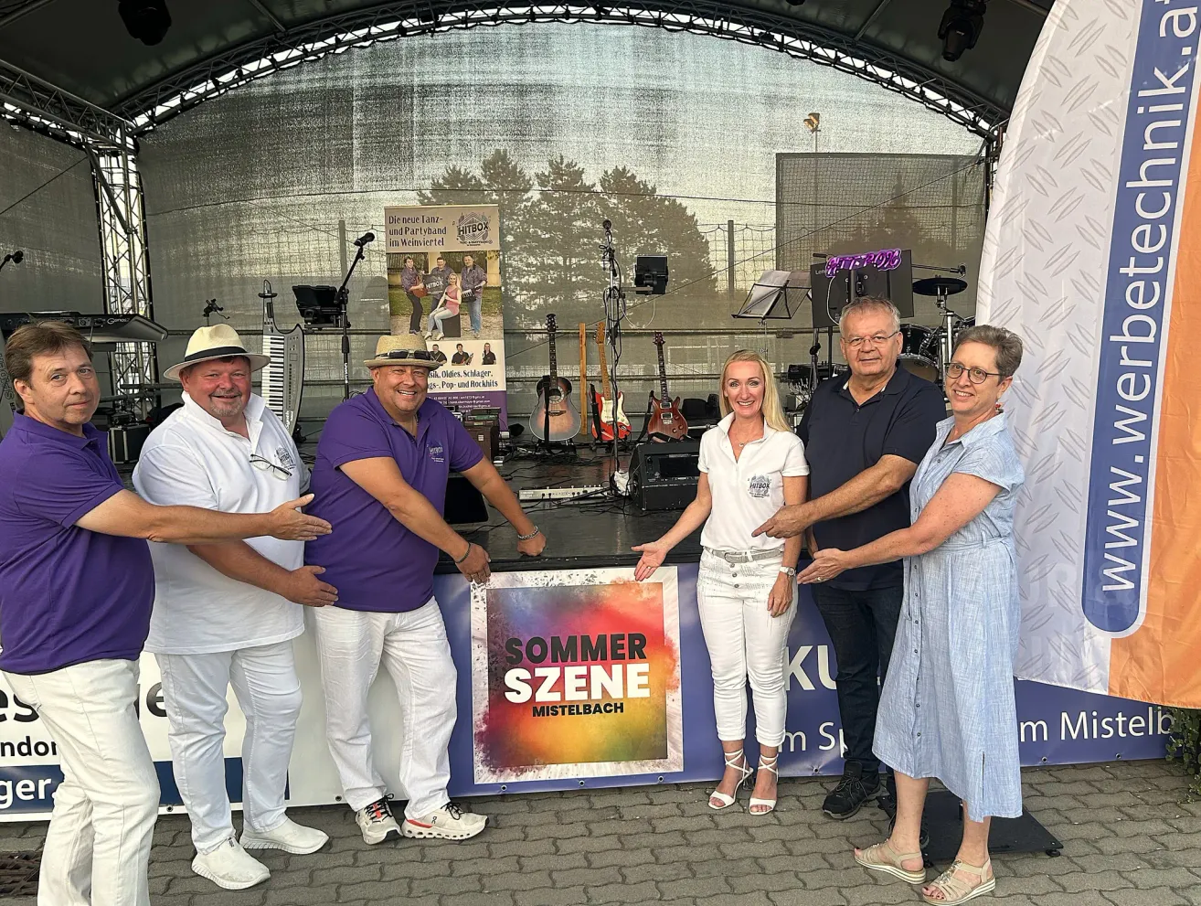 Fünf Personen stehen vor einer Bühne und lächeln für ein Foto. Eine Frau in der Mitte hält ein Schild mit der Aufschrift 'Sommerszene Mistelbach'. Hinter ihnen sind Musikinstrumente zu sehen, und ein Banner ist auf der Bühne angebracht.