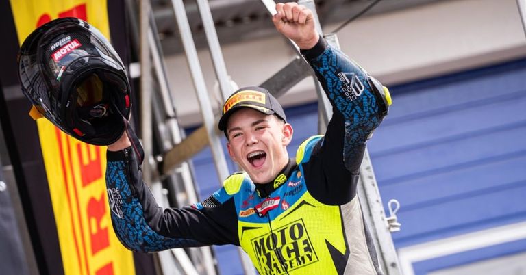 A young male motorcyclist celebrates a victory in a race, holding his helmet in the air with a big smile on his face. He wears a yellow and blue racing suit with the words 'MOTO RACE ACADEMY'.