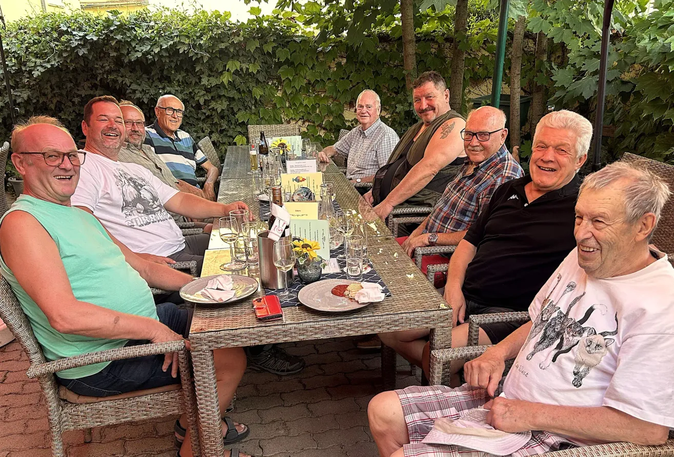 Eine Gruppe von Männern sitzt draußen an einem Tisch, lächelt und posiert für ein Foto. Der Tisch ist mit Tellern, Gläsern und einem Blumen-Zentrestück gedeckt.