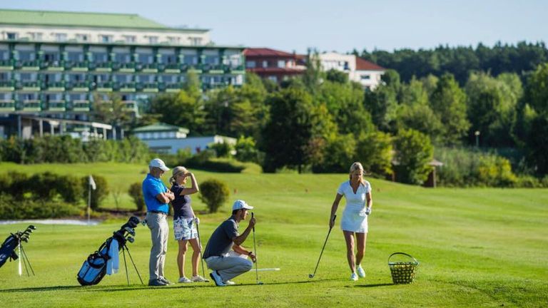 Vier Personen spielen Golf an einem sonnigen Tag. Ein Mann kniet und hält einen Golfschläger. Eine Frau geht auf einen Korb mit Golfbällen zu. Eine andere Frau steht und schaut nach oben. Ein Mann steht und schaut die Frau an.