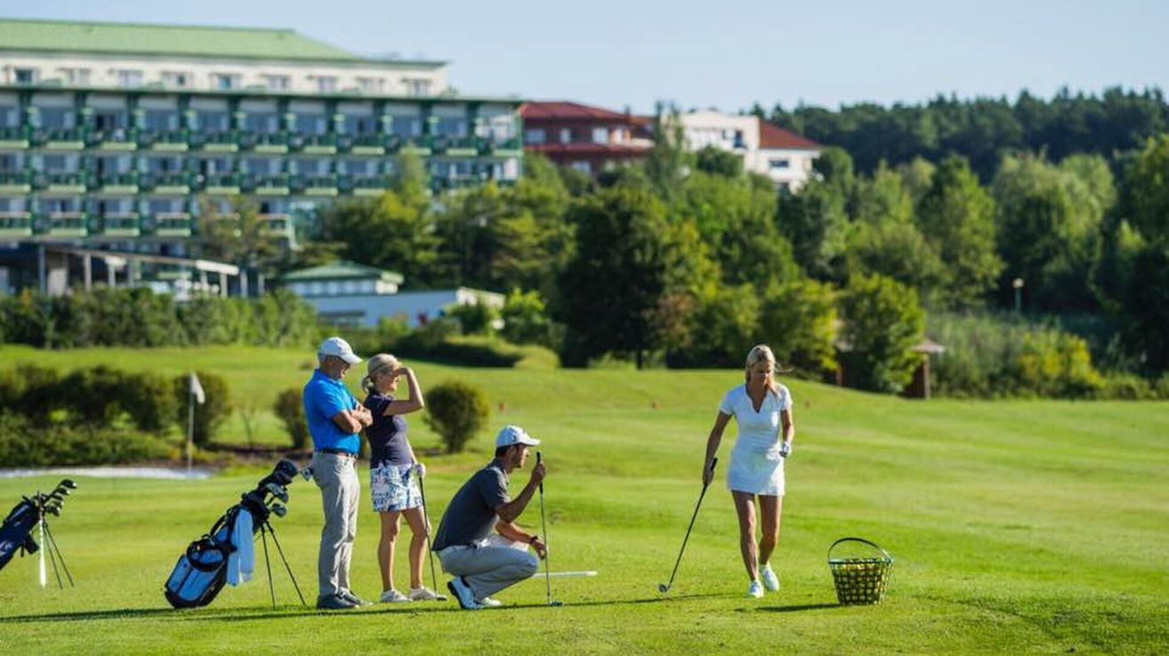 Vier Personen spielen Golf an einem sonnigen Tag. Ein Mann kniet und hält einen Golfschläger. Eine Frau geht auf einen Korb mit Golfbällen zu. Eine andere Frau steht und schaut nach oben. Ein Mann steht und schaut die Frau an.