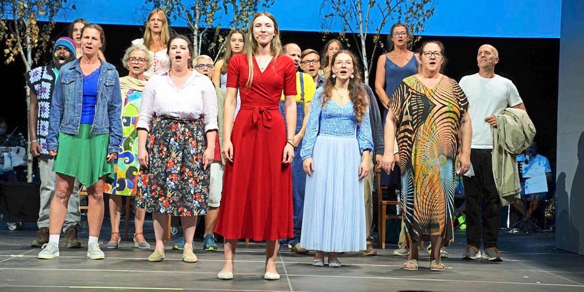 A group of people dressed in various colorful outfits are standing on a stage, possibly performing a musical number. They are all facing the same direction, with some singing and others watching. The stage is well-lit, and the background features trees and a blue wall.