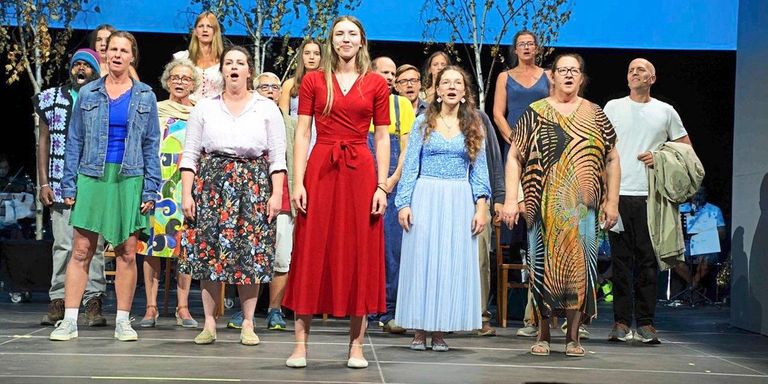 A group of people dressed in various colorful outfits are standing on a stage, possibly performing a musical number. They are all facing the same direction, with some singing and others watching. The stage is well-lit, and the background features trees and a blue wall.
