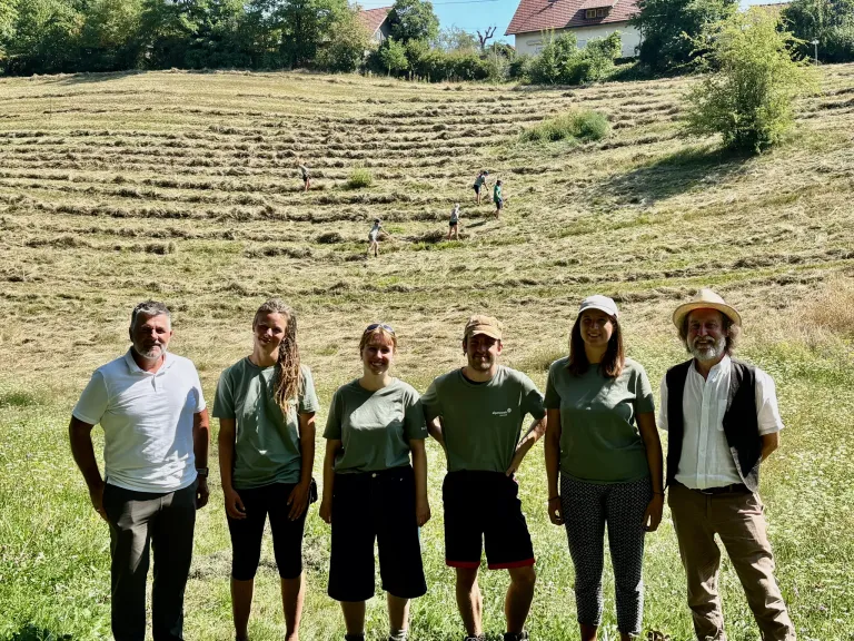 Eine Gruppe von Menschen in grünen Shirts steht auf einem Feld mit einigen Leuten in der Ferne. Das Feld ist von Bäumen und Häusern umgeben.