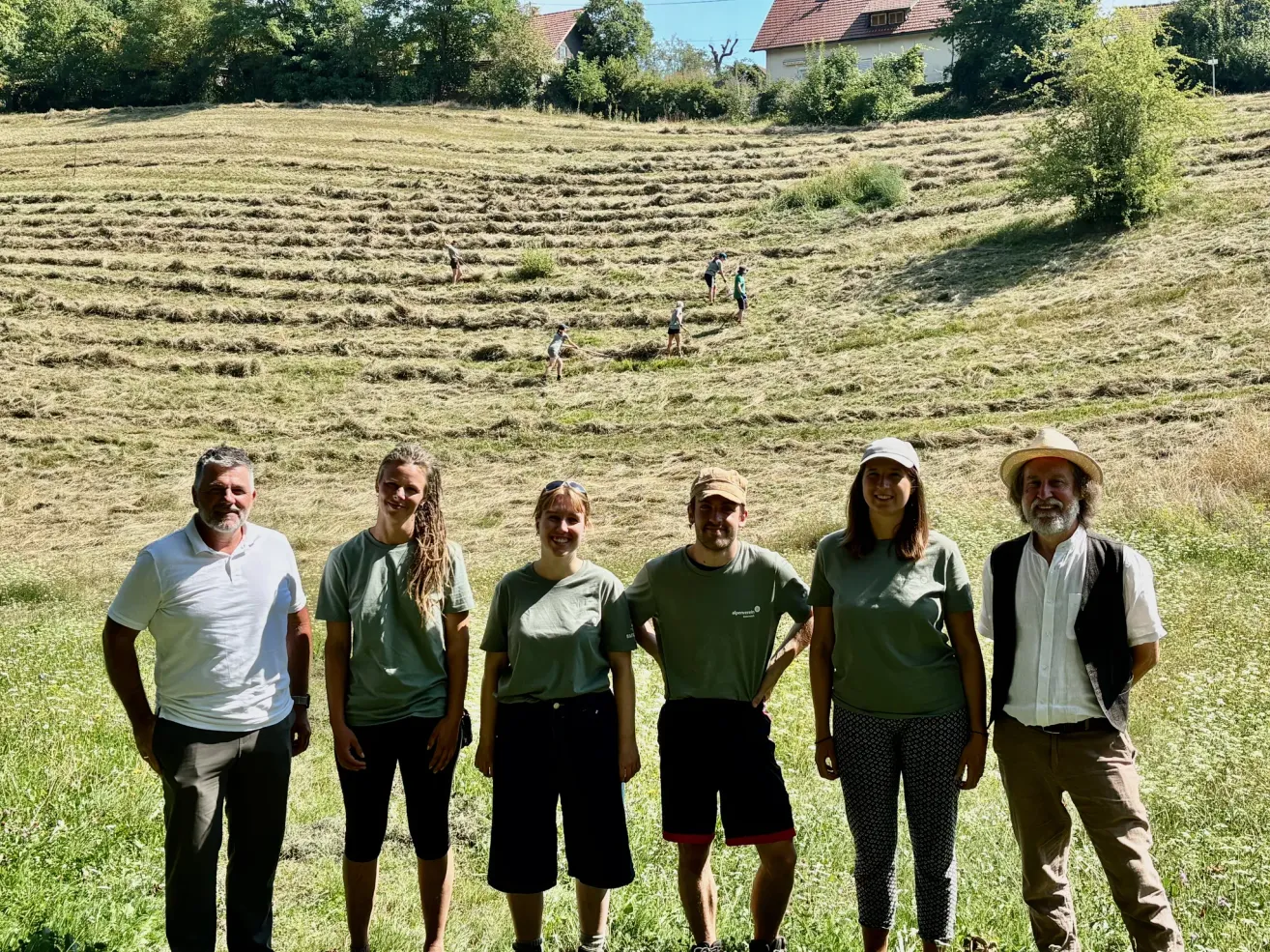 Eine Gruppe von Menschen in grünen Shirts steht auf einem Feld mit einigen Leuten in der Ferne. Das Feld ist von Bäumen und Häusern umgeben.