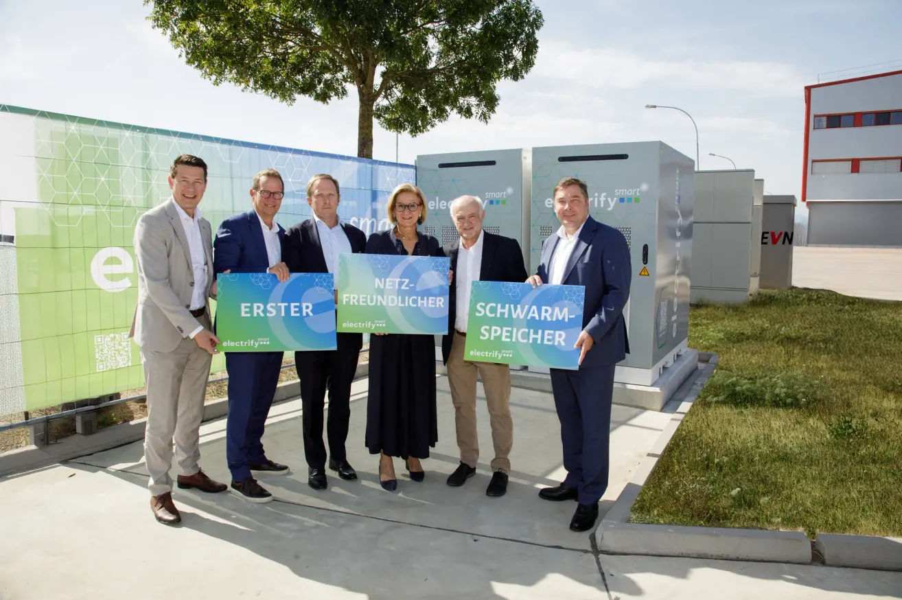 Five people stand in front of a large electrical storage unit, holding signs that read 'Netz-freundlicher' and 'Erster Schwarm-speicher'. They are dressed formally and appear to be posing for a photograph.