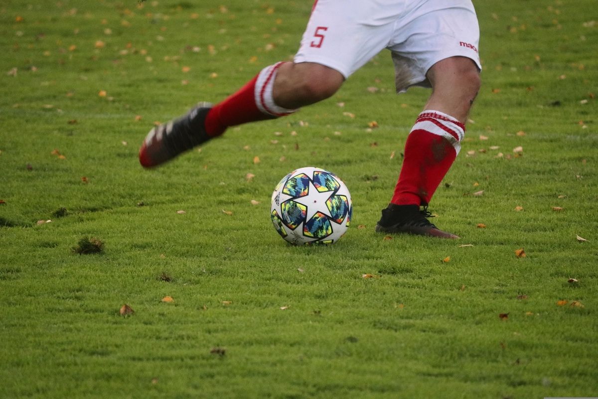 Ein Fußballspieler in einem weißen Trikot mit der Nummer 5 tritt einen Fußball mit einem farbenfrohen Design auf einem Rasenfeld.