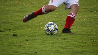 Ein Fußballspieler in einem weißen Trikot mit der Nummer 5 tritt einen Fußball mit einem farbenfrohen Design auf einem Rasenfeld.