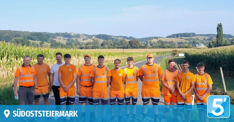 Eine Gruppe junger Männer in orangefarbenen Uniformen steht in einer Reihe auf einer Straße mit einem Maisfeld im Hintergrund.