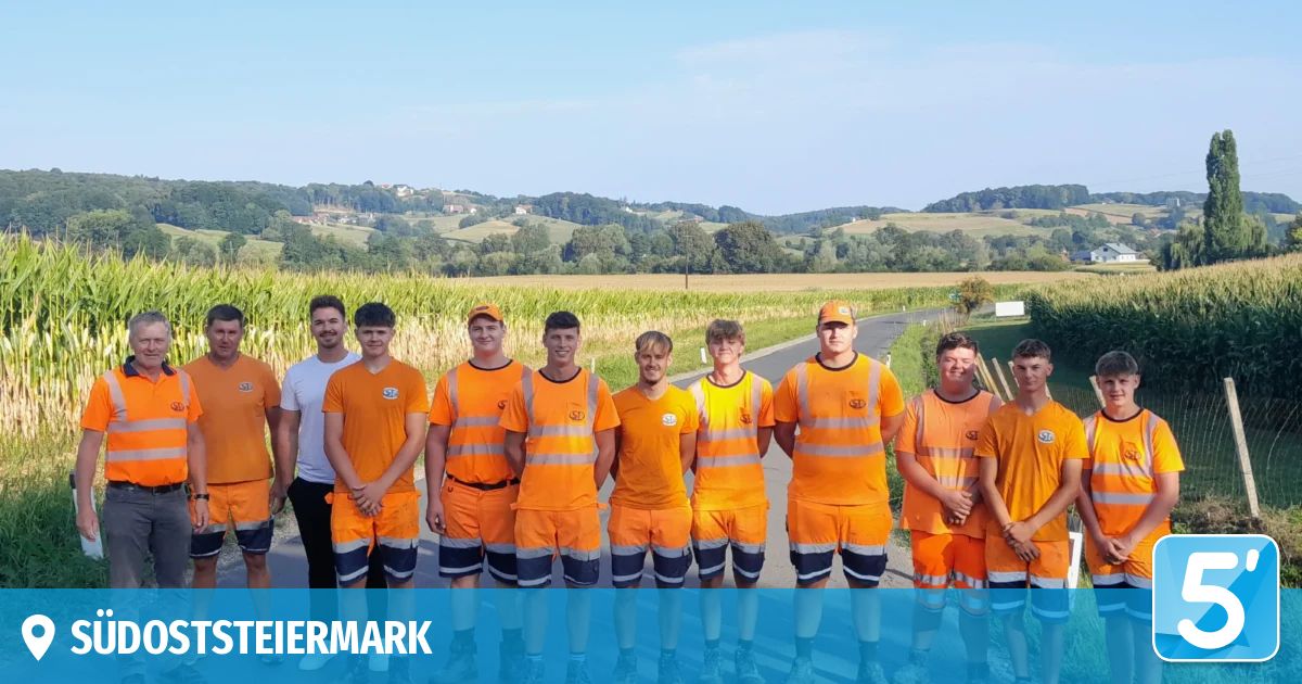 Eine Gruppe junger Männer in orangefarbenen Uniformen steht in einer Reihe auf einer Straße mit einem Maisfeld im Hintergrund.