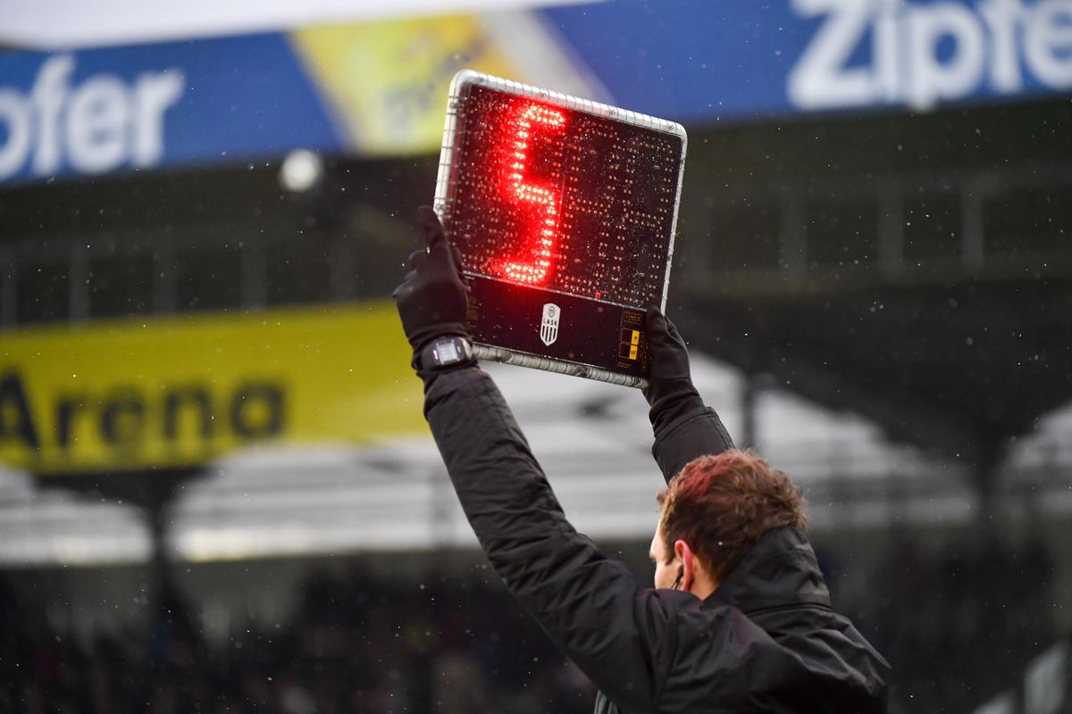 Ein Mann hält ein digitales Display mit der roten Zahl 5 hoch, vor einem verschwommenen Hintergrund mit Regen und einem gelben Banner.