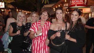 Vier Frauen lächeln und posieren für ein Foto bei einer Nachtveranstaltung. Sie halten Weingläser in der Hand. Die Frau in der Mitte trägt ein rot-weiß gestreiftes Kleid.
