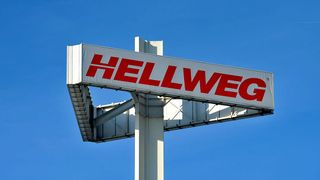 Ein großes weißes Schild mit roten Buchstaben, die 'HELLWEG' lesen, ist an einem weißen Pfosten gegen einen klaren blauen Himmel montiert.