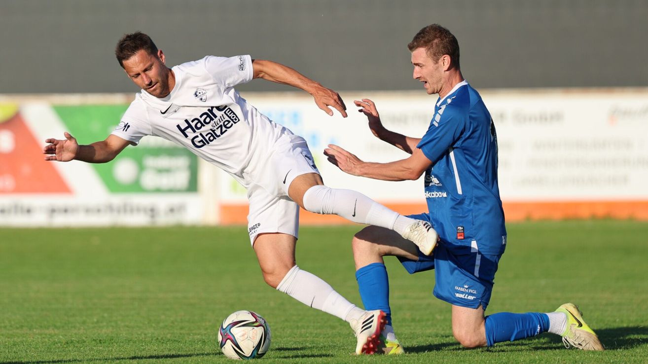 Zwei Fußballspieler sind auf einem Feld, einer in Weiß und der andere in Blau. Der Spieler in Weiß tritt den Ball, während der Spieler in Blau versucht, ihn zu blockieren.