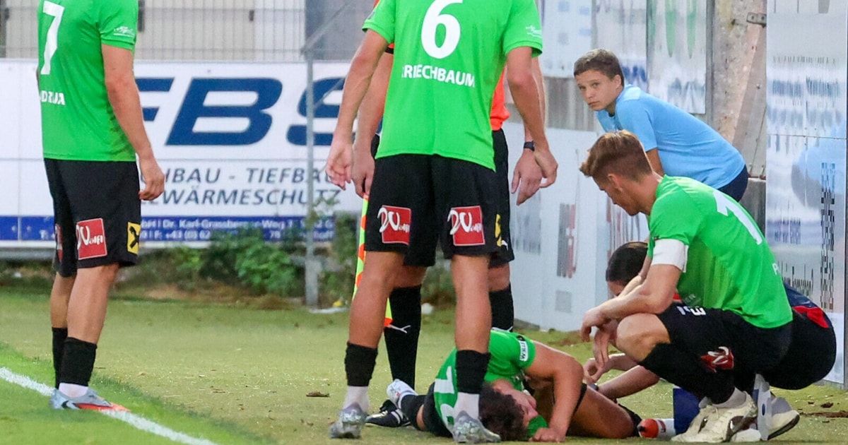 Fußballspieler auf einem Feld, einer liegt verletzt, ein anderer überprüft ihn, und zwei andere stehen dahinter, einer trägt ein grünes Trikot mit der Nummer 6.