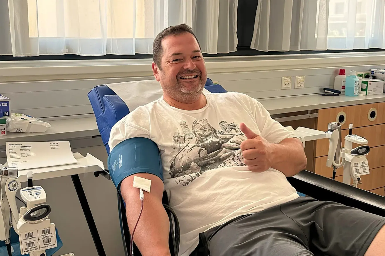 Ein Mann in einem Krankenhausstuhl, der ein weißes T-Shirt mit einem grafischen Design trägt, lächelt und gibt einen Daumen hoch. Er hat eine Blutdruckmanschette am linken Arm.